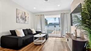 A modern living room with a black sofa, a white coffee table, and a wooden floor. There is a large window with curtains, a TV on a wooden cabinet, and a plant on the right. - Swaying Palms Unit 1: 2 BR 2 Bath Retreat, Near Beach, Heated Pool, Free Parking (Fort Lauderdale)
