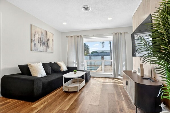 A modern living room with a black sofa, a white coffee table, and a wooden floor. There is a large window with curtains, a TV on a wooden cabinet, and a plant on the right. - Swaying Palms Unit 1: 2 BR 2 Bath Retreat, Near Beach, Heated Pool, Free Parking (Fort Lauderdale)
