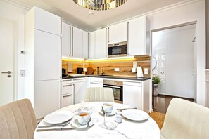 A modern kitchen with white cabinetry, a built-in oven, and a dining area with a marble table and chairs. - Gleam Homes Art Deco Dresden HafenCity (Dresden)