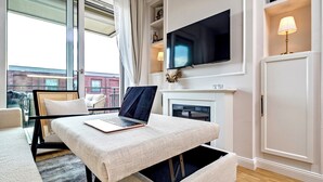 Una sala moderna con una laptop sobre un otomán texturizado, un televisor de pantalla plana sobre una chimenea y un amplio ventanal con cortinas.