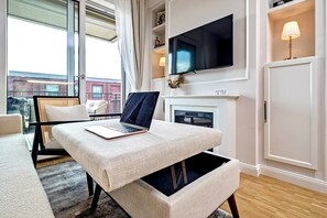 A modern living room with a laptop on a textured ottoman, a flat-screen TV above a fireplace, and a large window with curtains. - Gleam Homes Art Deco Dresden HafenCity (Dresden)