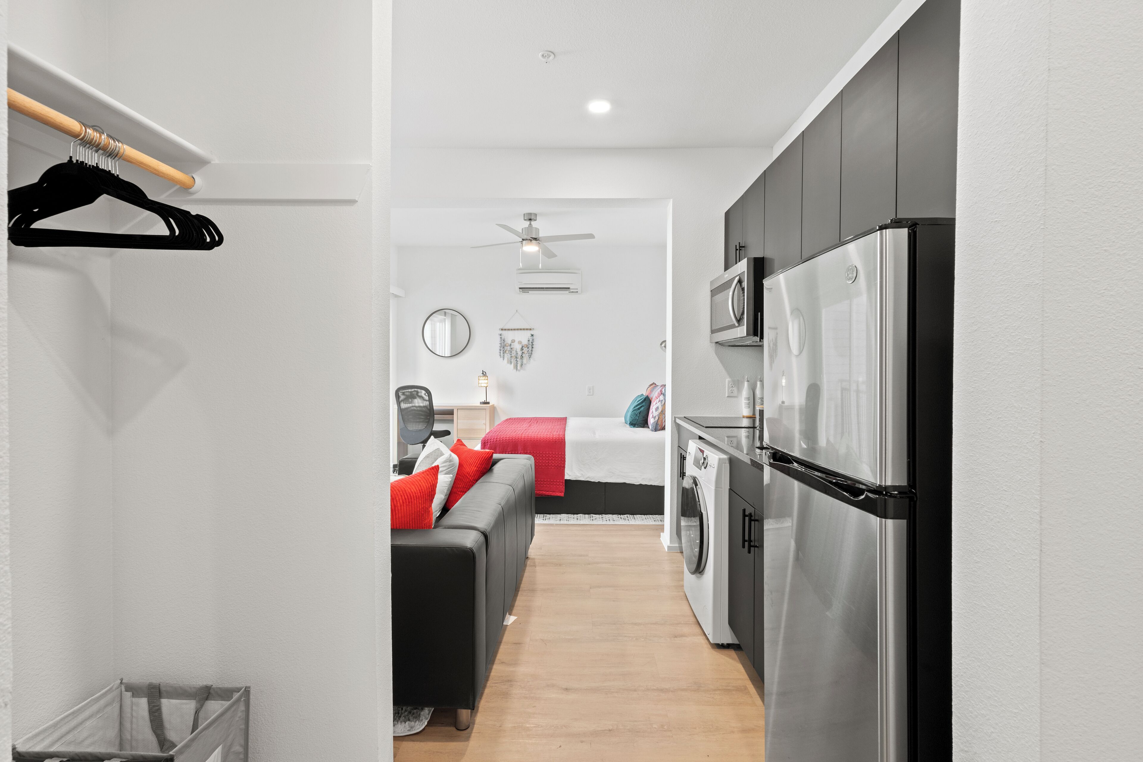 A compact hotel room with a kitchenette, a bed with red bedding, a black sofa, and a ceiling fan.