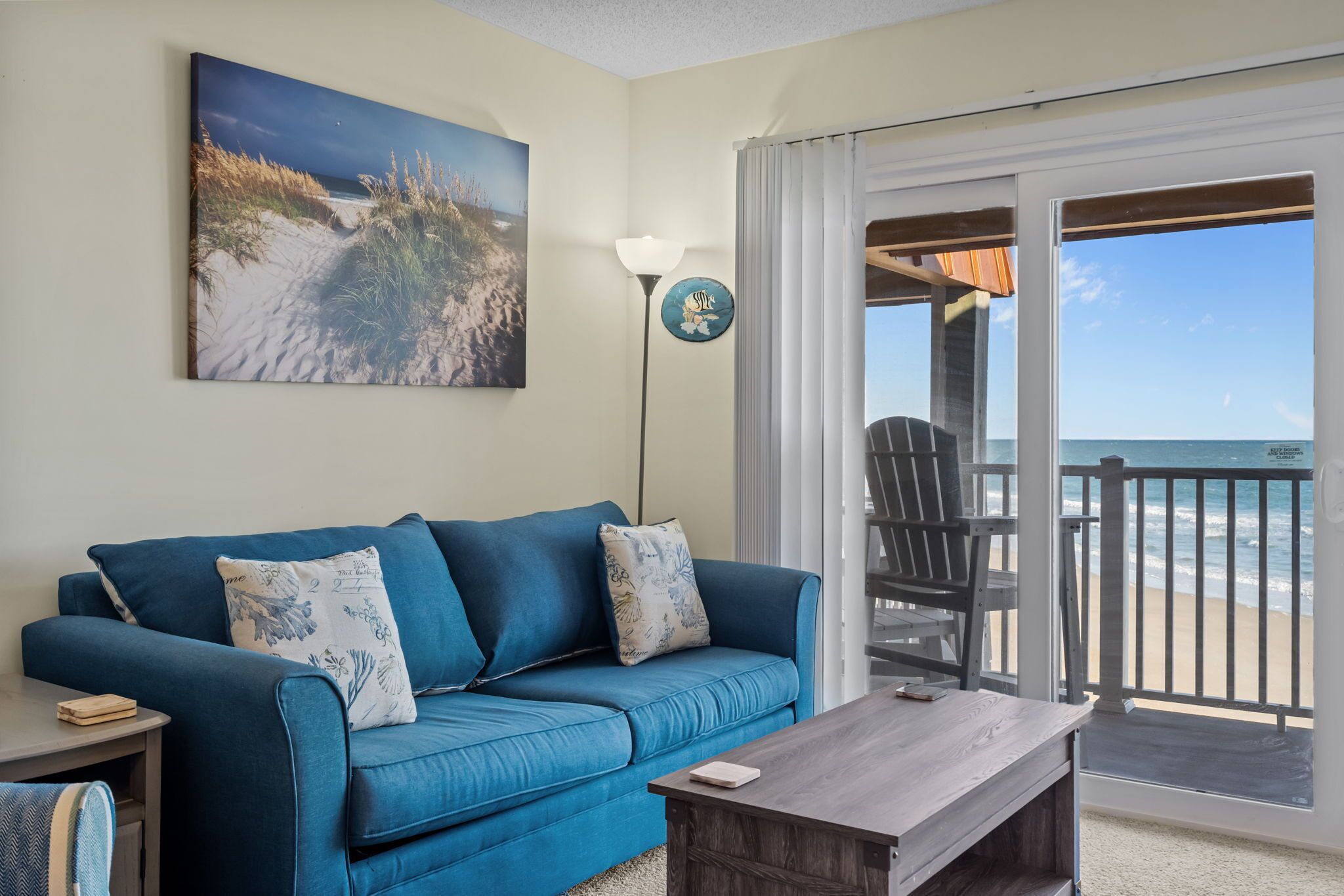 Ein Wohnzimmer mit einem blauen Sofa, einem Holztisch, einem Gemälde von einem Strand, einer Stehlampe und Blick auf den Strand durch eine Schiebetür.