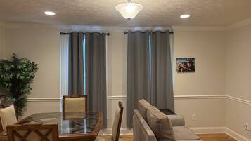 A living room with a glass-top dining table, cushioned chairs, a sofa, and a ceiling fan.