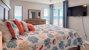 Una camera da letto con un letto, una ventola a soffitto, un comò, una TV e vista sull'oceano attraverso ampie finestre.