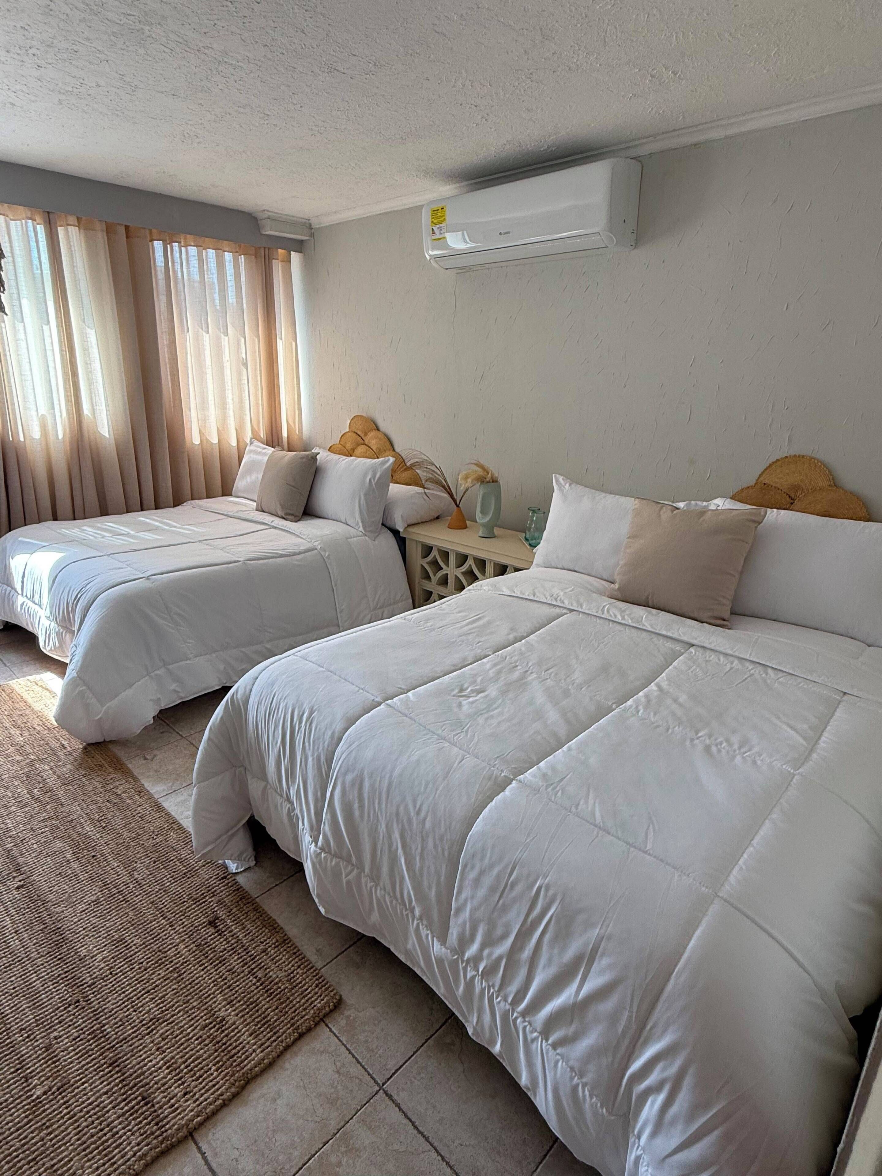 A hotel room with two beds, a small bedside table, a wall-mounted air conditioner, and a window with curtains.