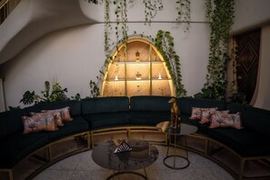 Een gebogen groene bank met gekleurde kussens, een glazen bijzettafeltje en een decoratief hondenbeeld.