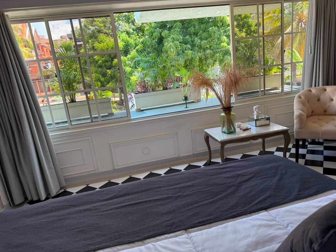 Une chambre à coucher avec un lit, une petite table sur laquelle est posé un vase, une chaise et une fenêtre donnant sur un paysage verdoyant et des bâtiments.