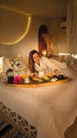 Una persona recostada en una cama con una bandeja de desayuno, un muro decorativo y una planta colgante.