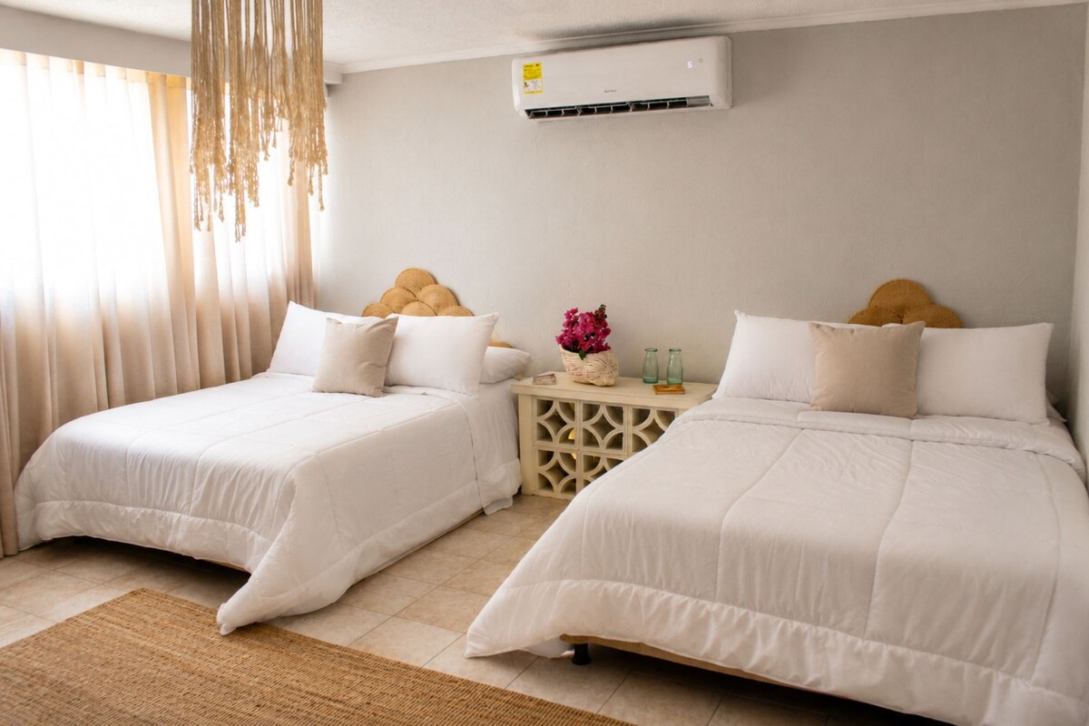 A hotel room with two beds, a small table with a vase of flowers, and an air conditioning unit on the wall.