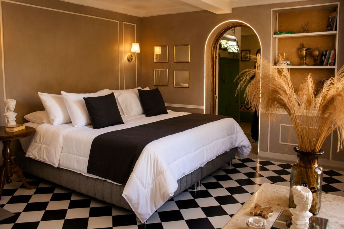 Une chambre d’hôtel avec un sol à carreaux, un grand lit avec une literie blanche et noire, une table de chevet avec une lampe et un vase contenant des plantes séchées.