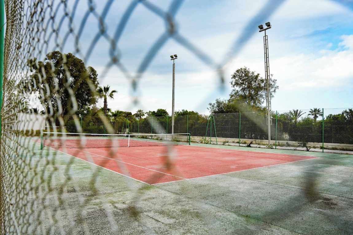 A tennis court with a chain-link fence, floodlights, and trees in the background.