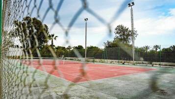 A tennis court with a chain-link fence, floodlights, and trees in the background.