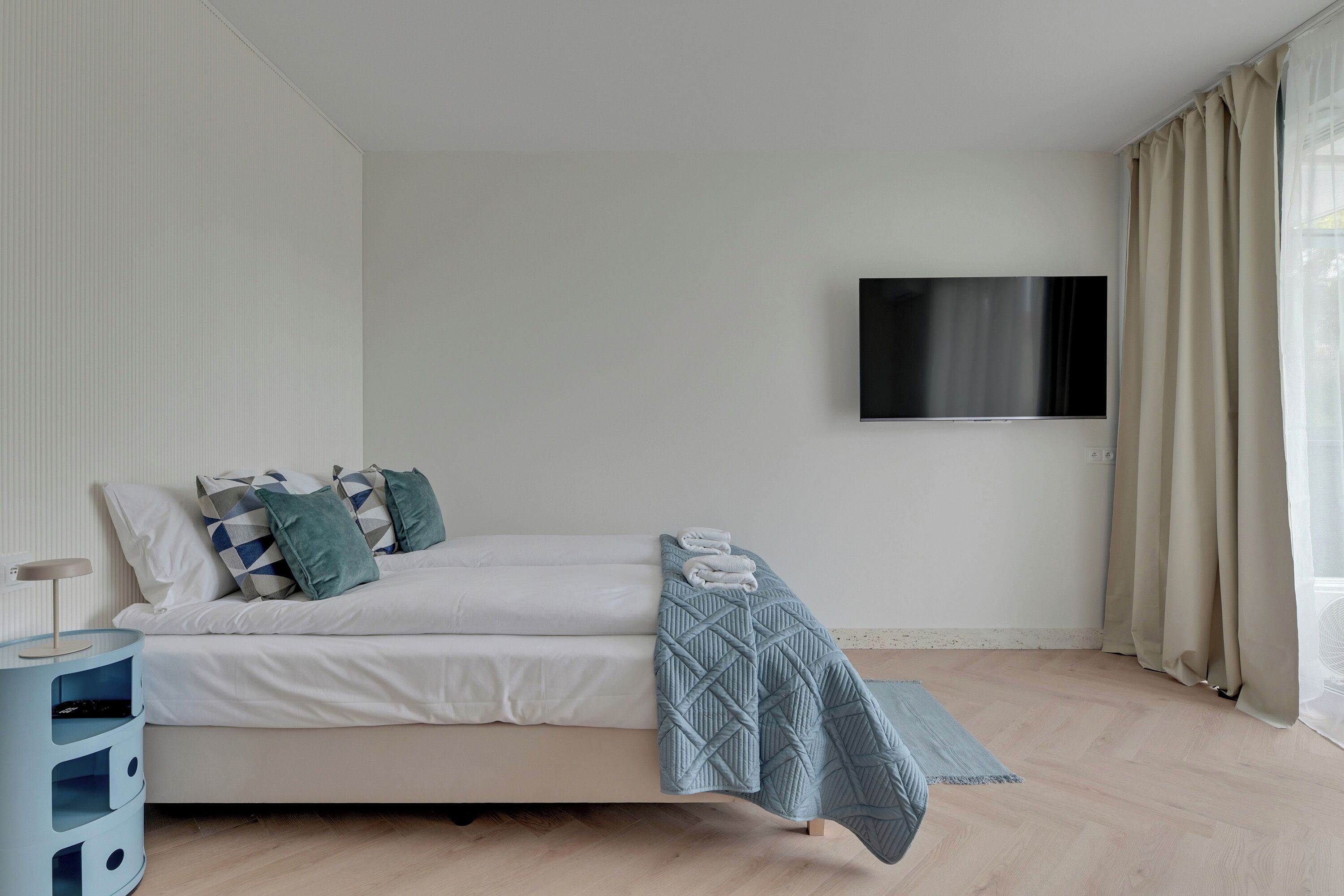A bedroom with a bed, a TV mounted on the wall, a bedside table, and a window with curtains.