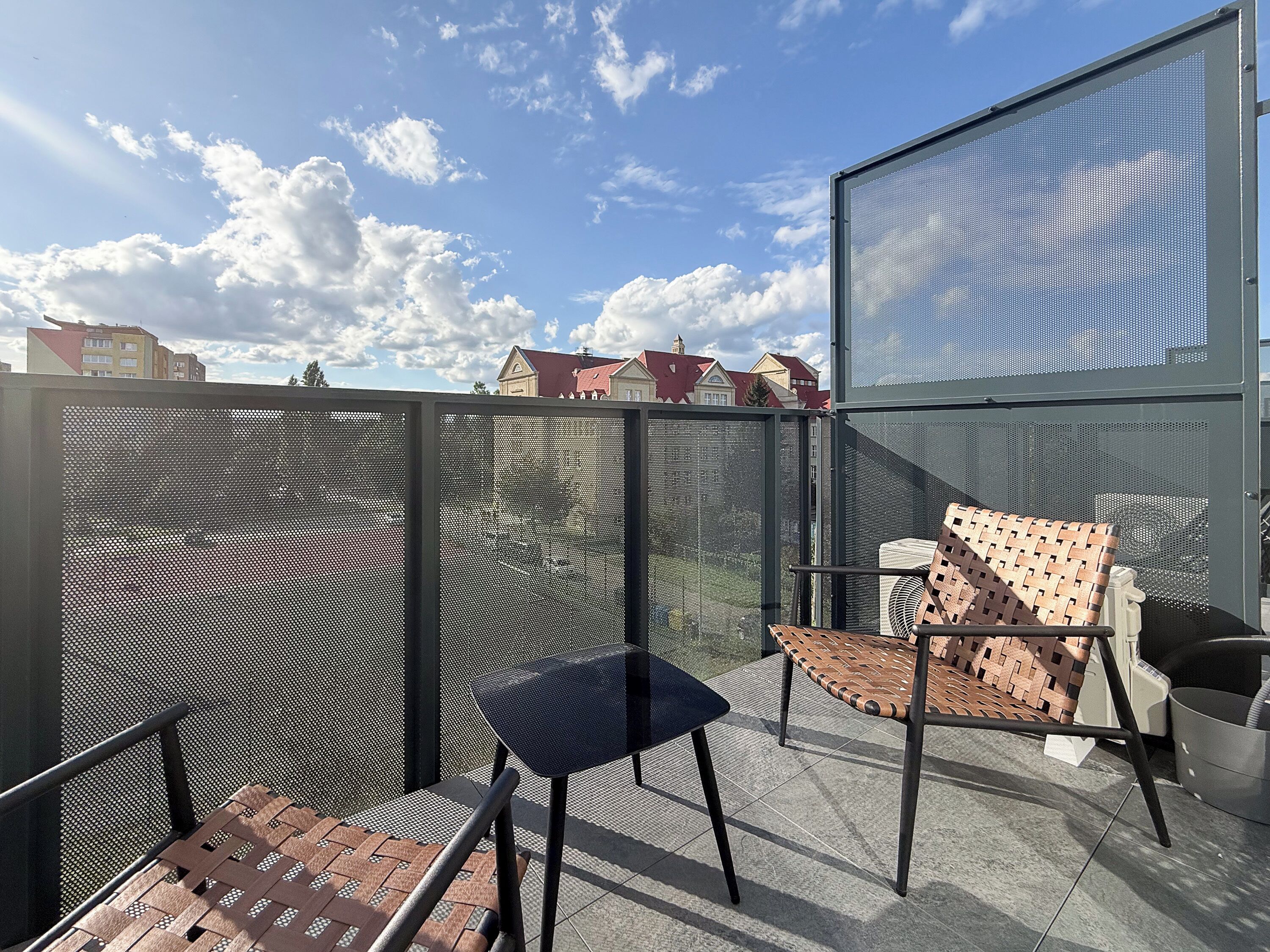 Uma varanda com duas cadeiras, uma com assento e encosto trançados, e outra com encosto firme. Uma grade de metal separa a varanda do interior do edifício.