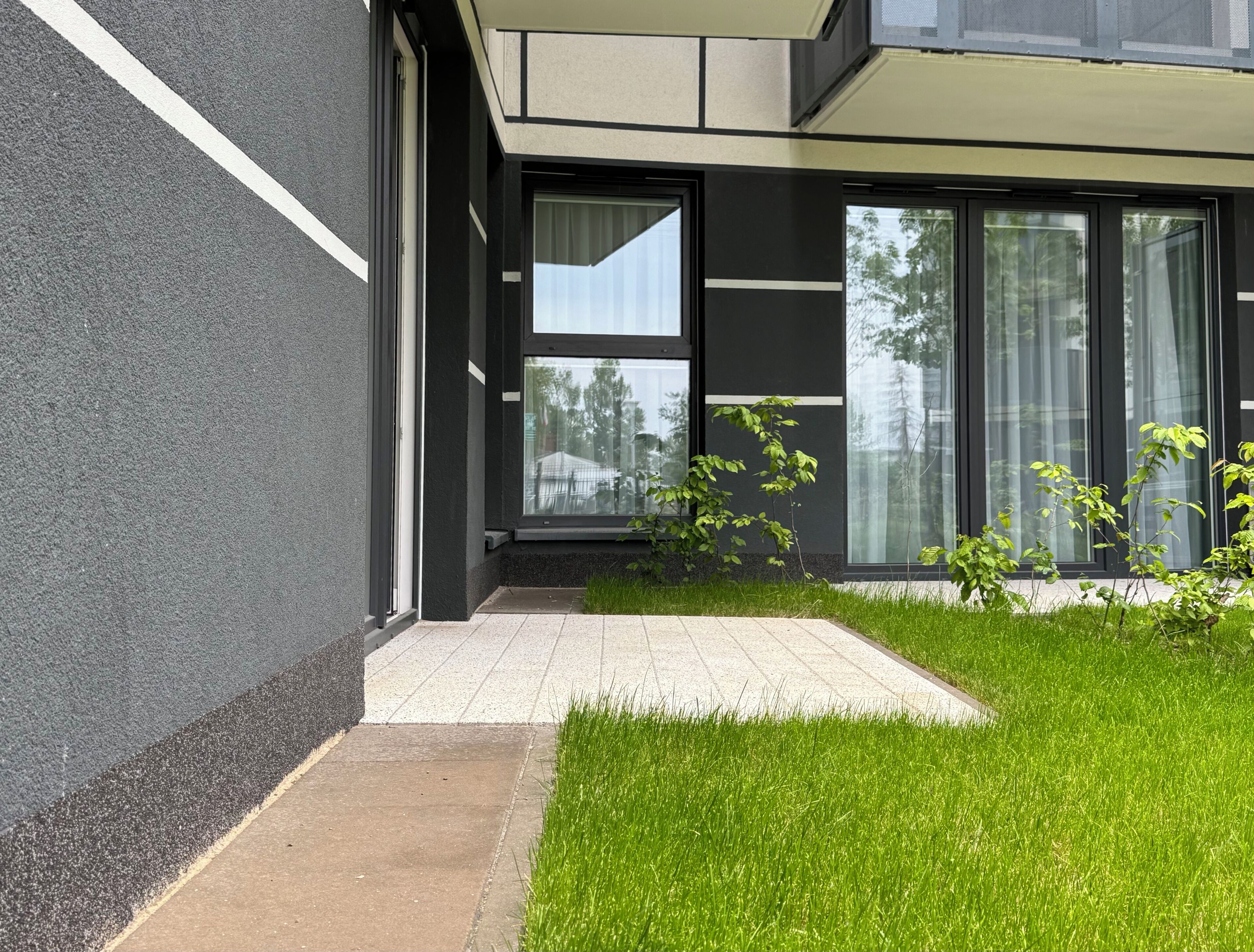 A modern building with a paved walkway, a small grassy area, and a window with a view of trees.