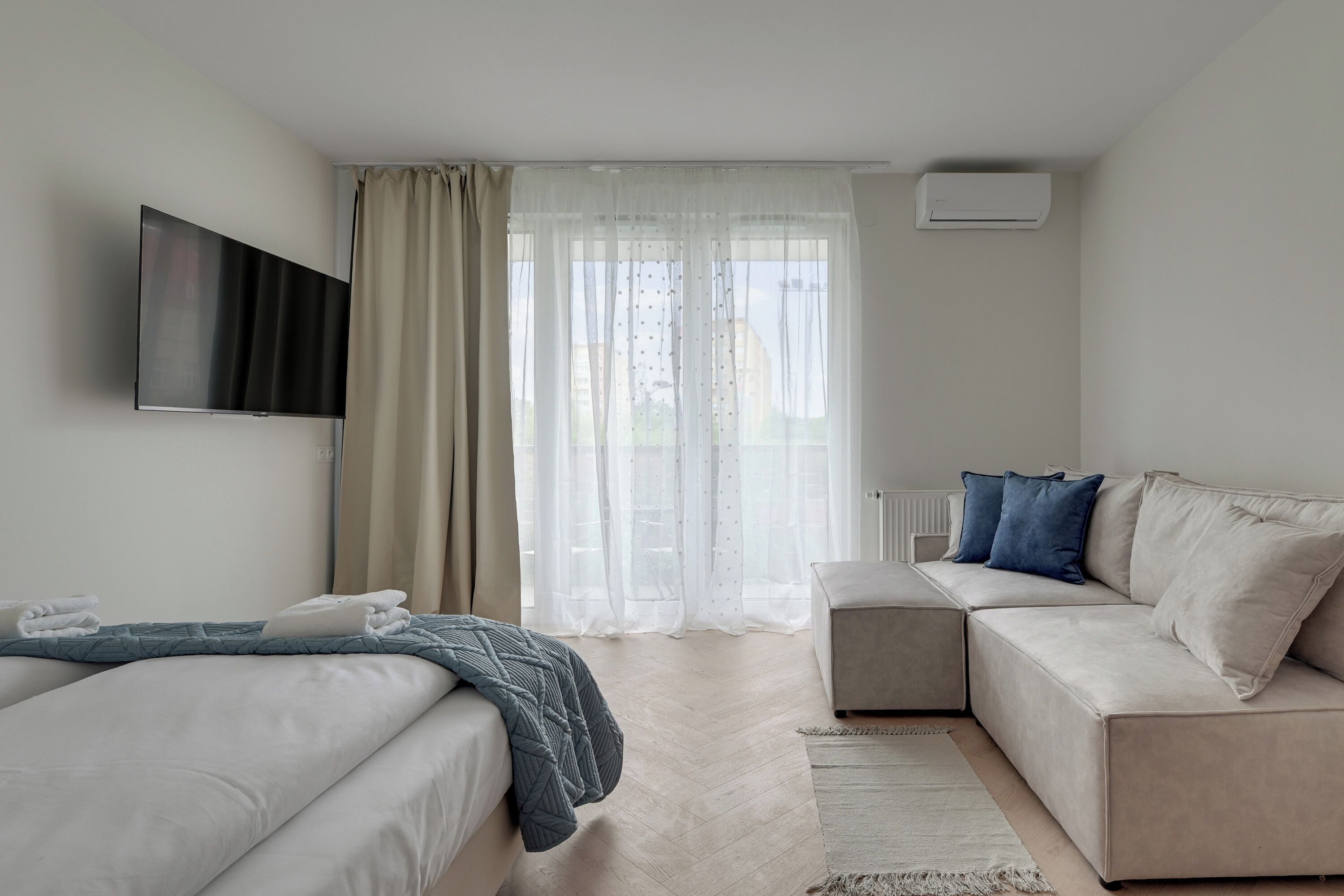 A modern hotel room with a bed, a sofa, a TV, and a window with curtains.