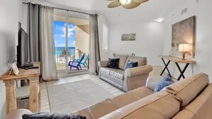 Sala de estar com sofá bege, uma mesa de centro e uma TV. Possui varanda com vista para o oceano.