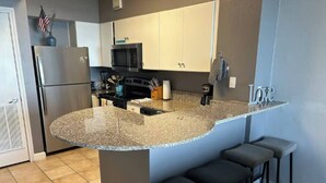 Cozinha com bancada de granito, geladeira, micro-ondas e pia. Há três banquetas de bar e um armário acima da bancada.