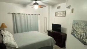 Quarto com cama, ventilador de teto, televisão, mesinha e um quadro na parede.
