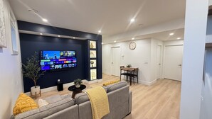 A modern living room with a flat-screen TV mounted on a dark wall, a grey sofa with yellow cushions, a small wooden table, and a dining area with a wooden table and chairs. - Modern 1BR Private Suite Near Airport and Amazing Shopping!Free Netflix, Parking (Calgary)