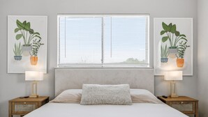 Une chambre à coucher avec un lit, deux tables de chevet avec des lampes, une fenêtre avec des stores et deux illustrations de plantes encadrées accrochées au mur.
