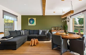 A modern living room with a sectional sofa, a round wooden dining table, and a large window providing a view of the outdoors.