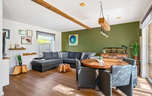 A modern living room with a sectional sofa, a round wooden dining table, and a large window providing natural light.