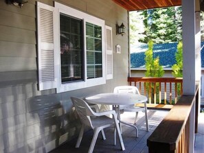 A covered patio with a table and chairs, white shutters, and a view of greenery. - Lile II, West in Lake Almanor West, CA (Lake Almanor West)