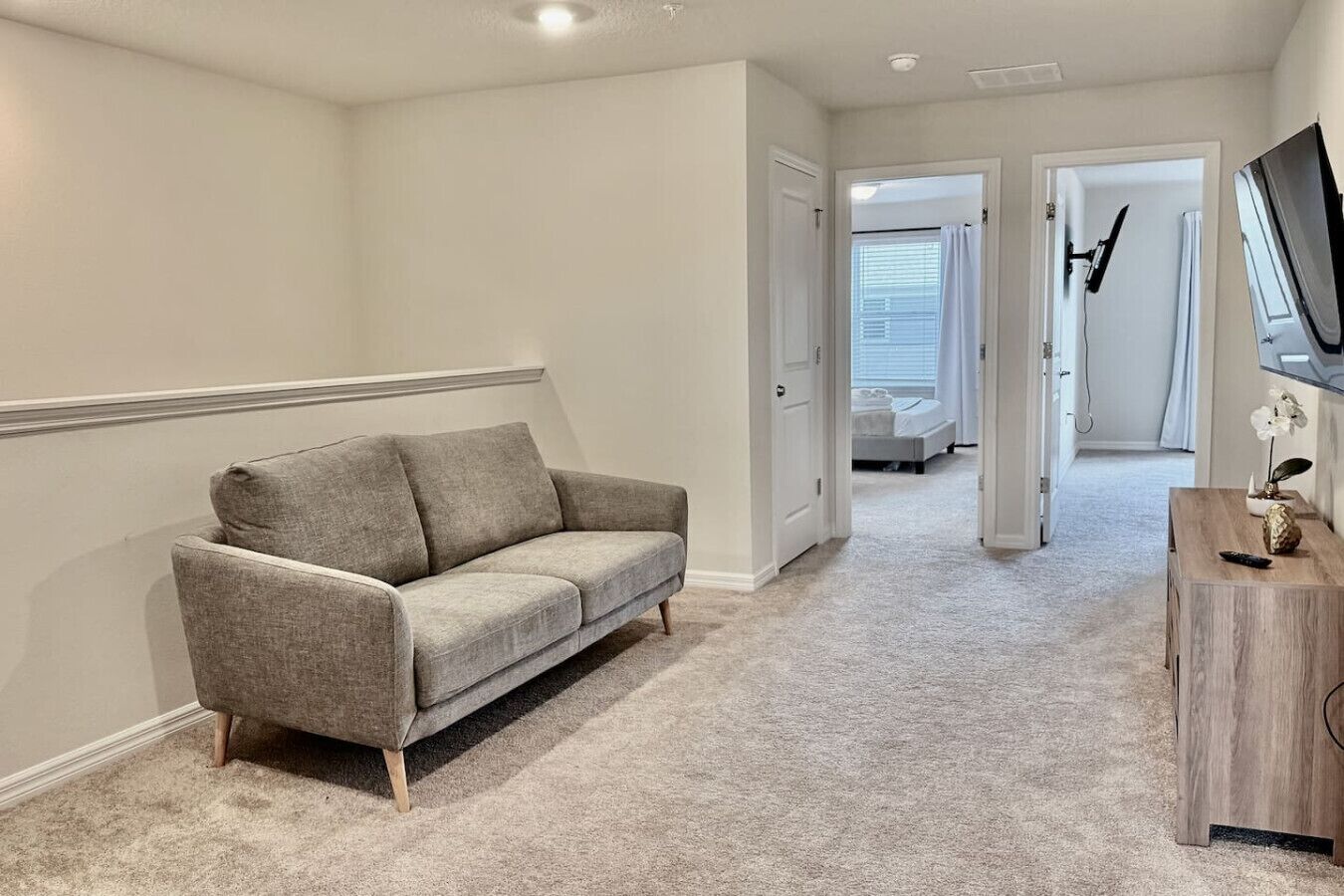 Un couloir avec un canapé gris, une table d’appoint en bois et un téléviseur fixé au mur.
