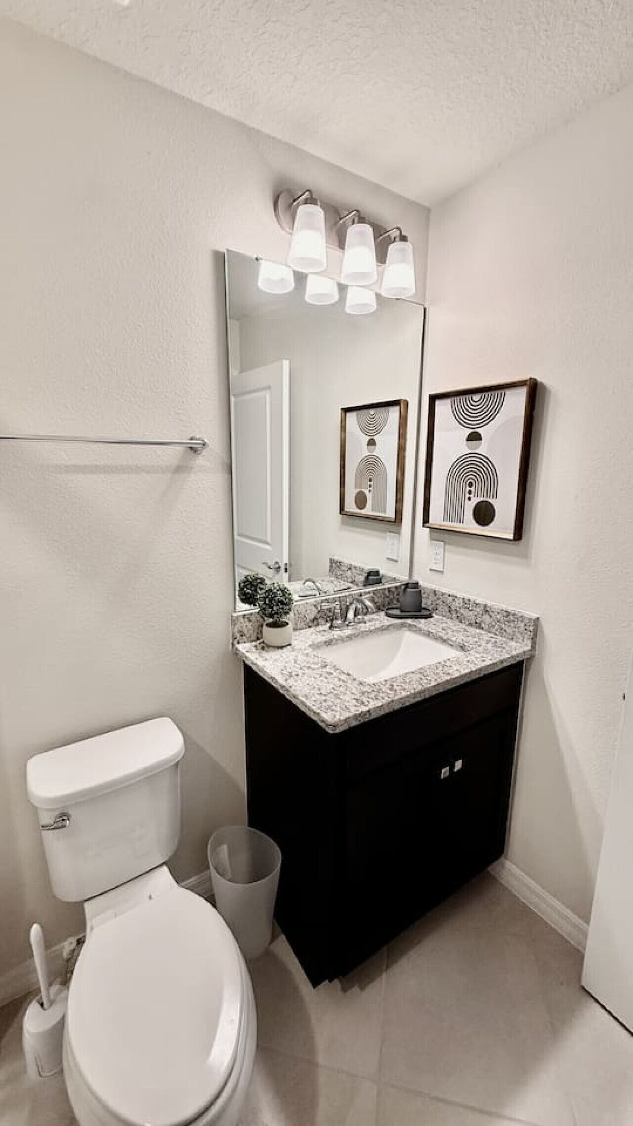 Une salle de bain moderne avec des toilettes blanches, un lavabo doté d’un plan de travail en marbre et un meuble-lavabo noir. Deux œuvres d’art encadrées sont accrochées au-dessus du miroir, et un porte-serviettes est fixé au mur.