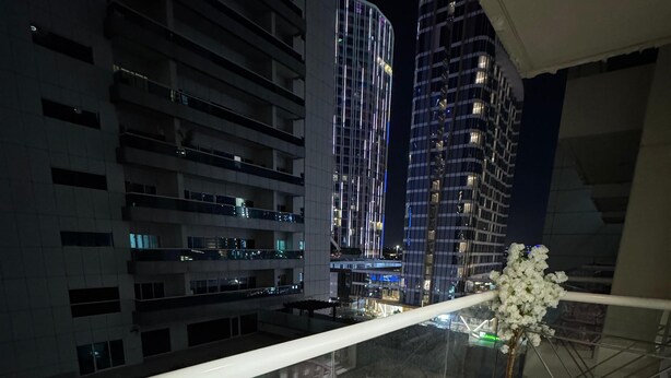 Ein Balkon mit rosafarbenen Stühlen und einem Glastisch, mit Blick auf eine nächtliche Skyline moderner Gebäude.