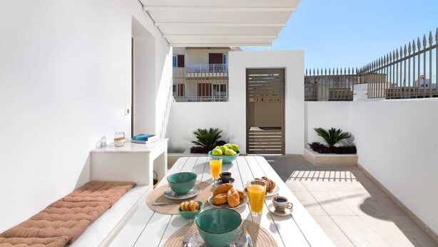 Ein sonniger Außenbereich mit Essplatz und weißem Tisch, gedeckt für ein Frühstück mit Croissants, Früchten und Orangensaft.