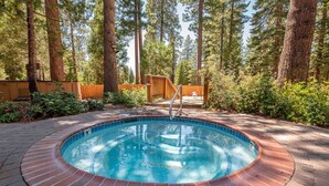 A circular hot tub with a brick border, surrounded by a wooden deck, set in a forested area. - Kings Beach Studio • Pool, Hot Tub, SUP, & Sauna • Kings Beach & Northstar (Tahoe Vista)