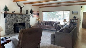 A cozy living room with a stone fireplace, wooden beams, a sofa set, and a bookshelf. - The Emerald Lake House (Lake Almanor)