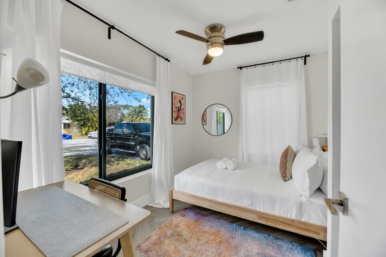 A bedroom with a bed, a desk, a ceiling fan, a window with curtains, and a view of the outside.