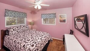 Un dormitorio con cama, escritorio, silla, ventilador de techo y dos ventanas con adornos.
