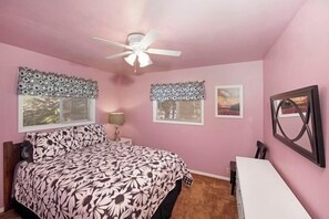 Ein Schlafzimmer mit einem Bett, einem Schreibtisch, einem Sessel, einem Deckenventilator und zwei Fenstern mit Zierrollvorhängen.