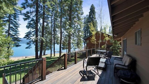 Terraza de madera con mobiliario exterior, con vista a un lago rodeado de altos árboles.