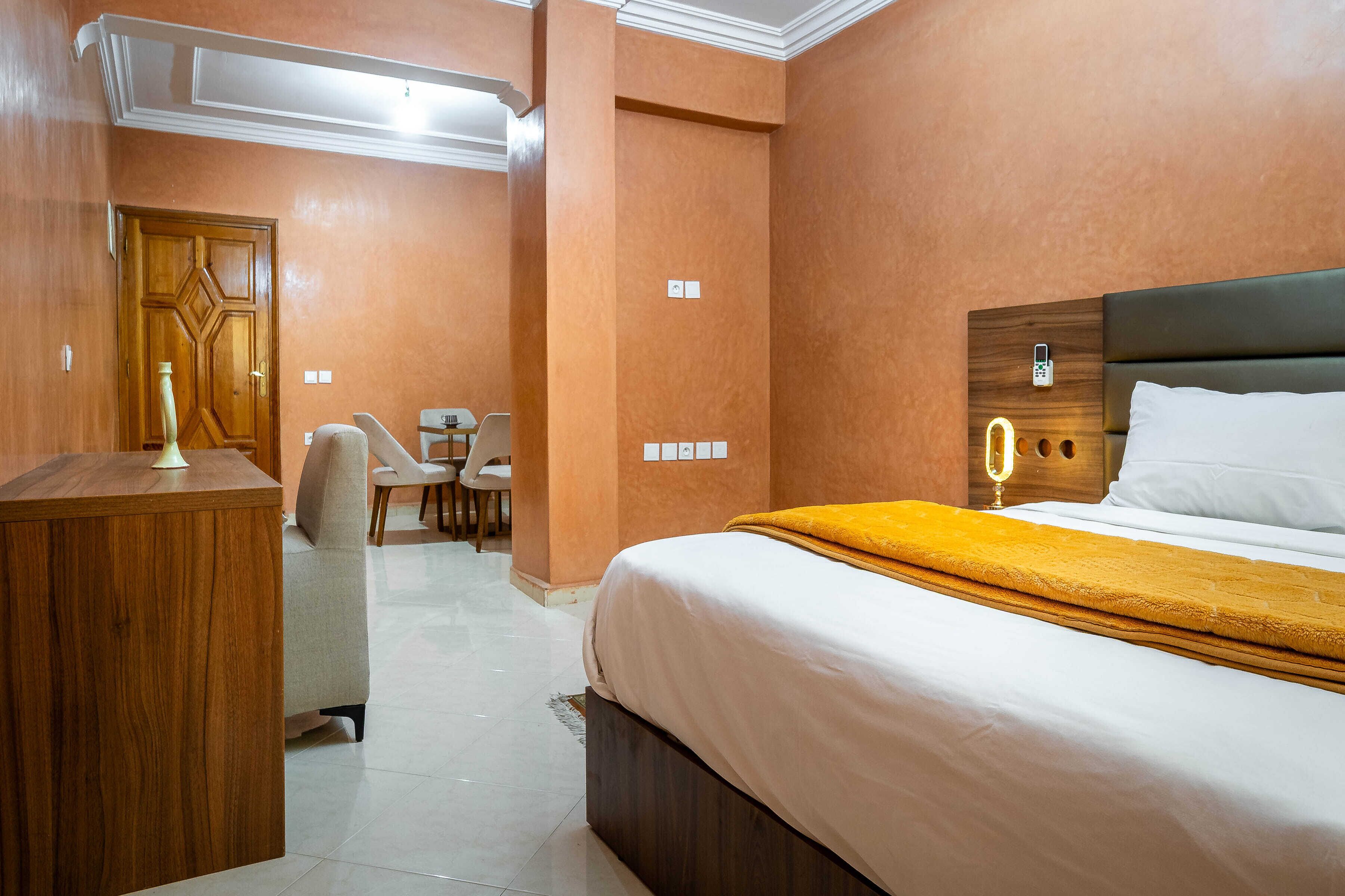 A hotel room with a wooden desk, a bed with a white and yellow bedspread, a wooden headboard, and a small table with chairs.