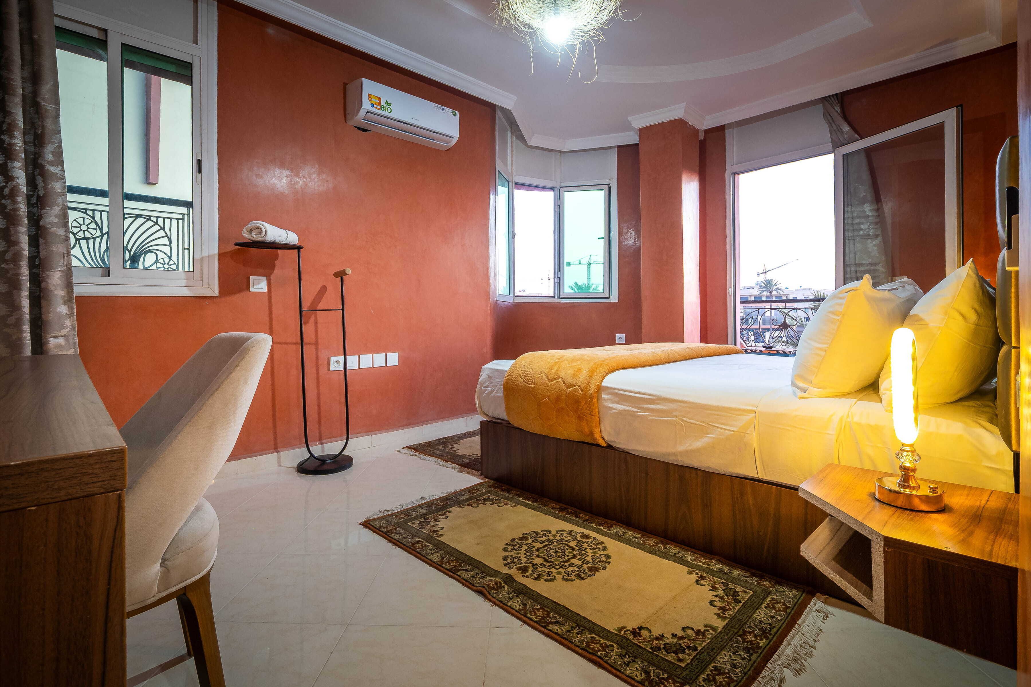 Une chambre d’hôtel comprenant un lit, une table de chevet avec une lampe, une chaise, un lampadaire, une fenêtre et un appareil de climatisation.