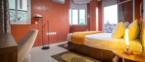 A hotel room with a bed, bedside table with a lamp, a chair, a standing lamp, a window, and an air conditioning unit.
