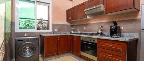 A compact kitchen with wooden cabinets, a built-in oven, a washing machine, and a window with a view of a balcony.