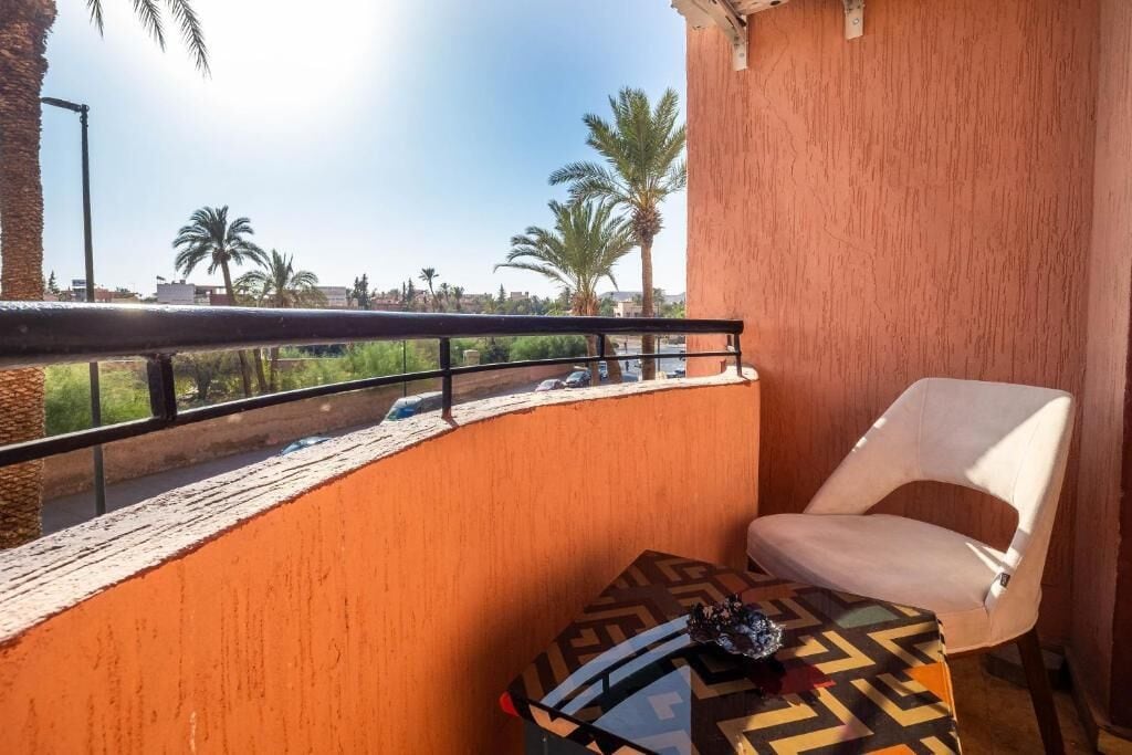 A balcony with a white chair, a patterned table, and a view of palm trees and a road.