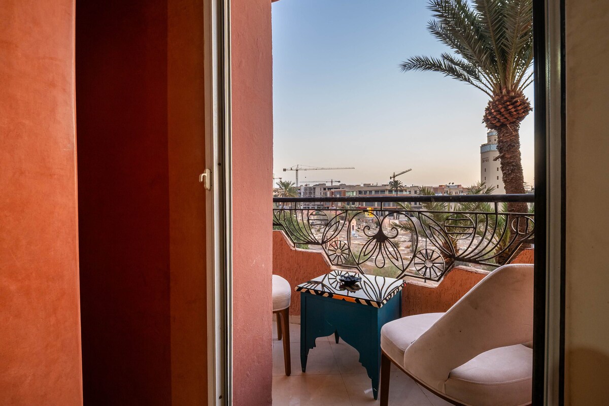 Un balcon avec une table et des chaises, un palmier, et une vue sur des immeubles et des grues de construction.