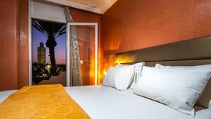 A hotel room with a bed, a view of a cityscape at dusk, and a palm tree outside the window.