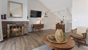 Una sala de estar con chimenea, un televisor montado en la pared y una zona de estar acogedora con un sillón y una mesa redonda.