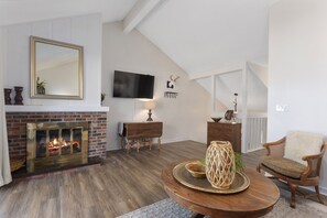 Ein Wohnzimmer mit Kamin, einem an der Wand befestigten Fernseher und einer gemütlichen Sitzecke mit Sessel und rundem Tisch.