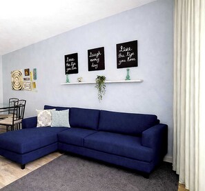 Ein modernes Wohnzimmer mit einem dunkelblauen Ecksofa, einem kleinen Esstisch mit Stühlen und drei motivierenden Wandbildern.