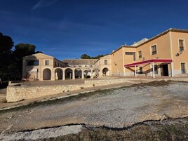 Un patio con un edificio que tiene tejado de baldosas y arcos, un muro de piedra y un toldo rojo.
